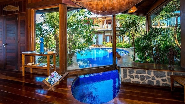 A wooden interior with a large window overlooking a swimming pool and lush greenery. - Sombra e Água Fresca Resort (Tibau do Sul)