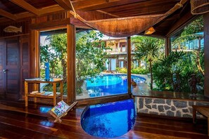 A wooden interior with a large window overlooking a swimming pool and lush greenery. - Sombra e Água Fresca Resort (Tibau do Sul)