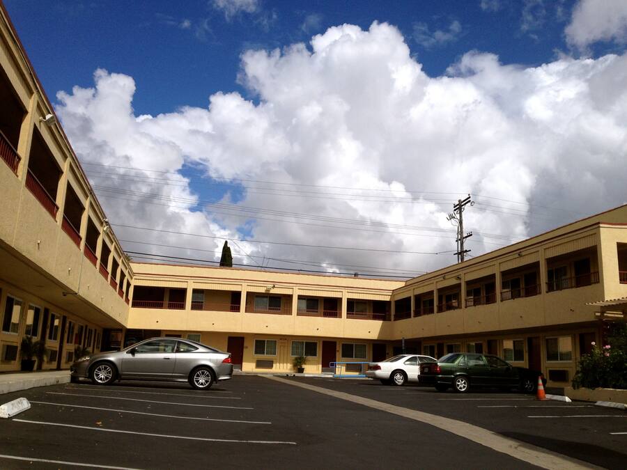 A parking lot with several cars, a building with multiple floors and balconies, and a clear sky with clouds.