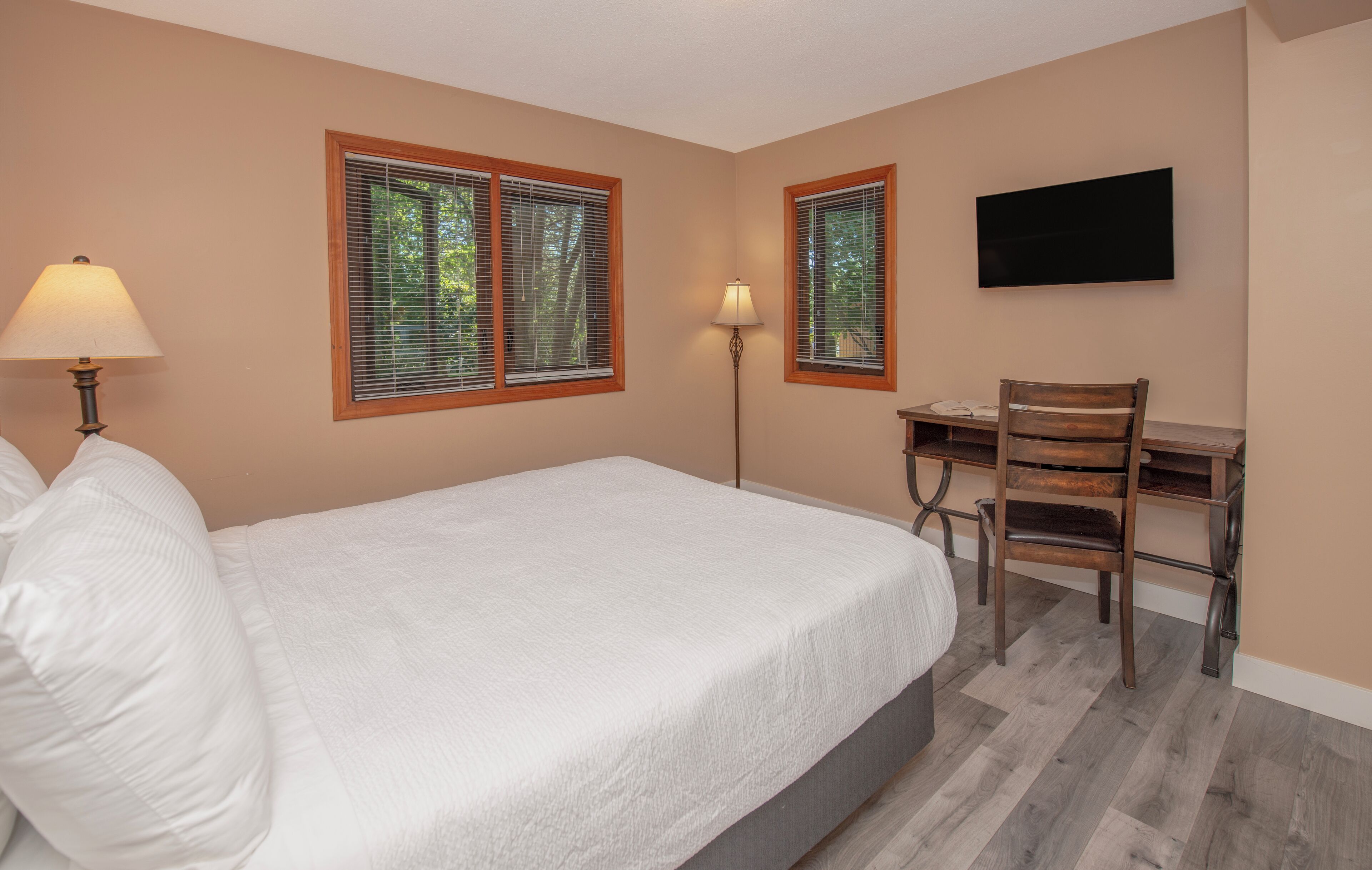 Une chambre à coucher avec un lit, deux fenêtres équipées de stores, un bureau avec une chaise et un téléviseur à écran plat fixé au mur.