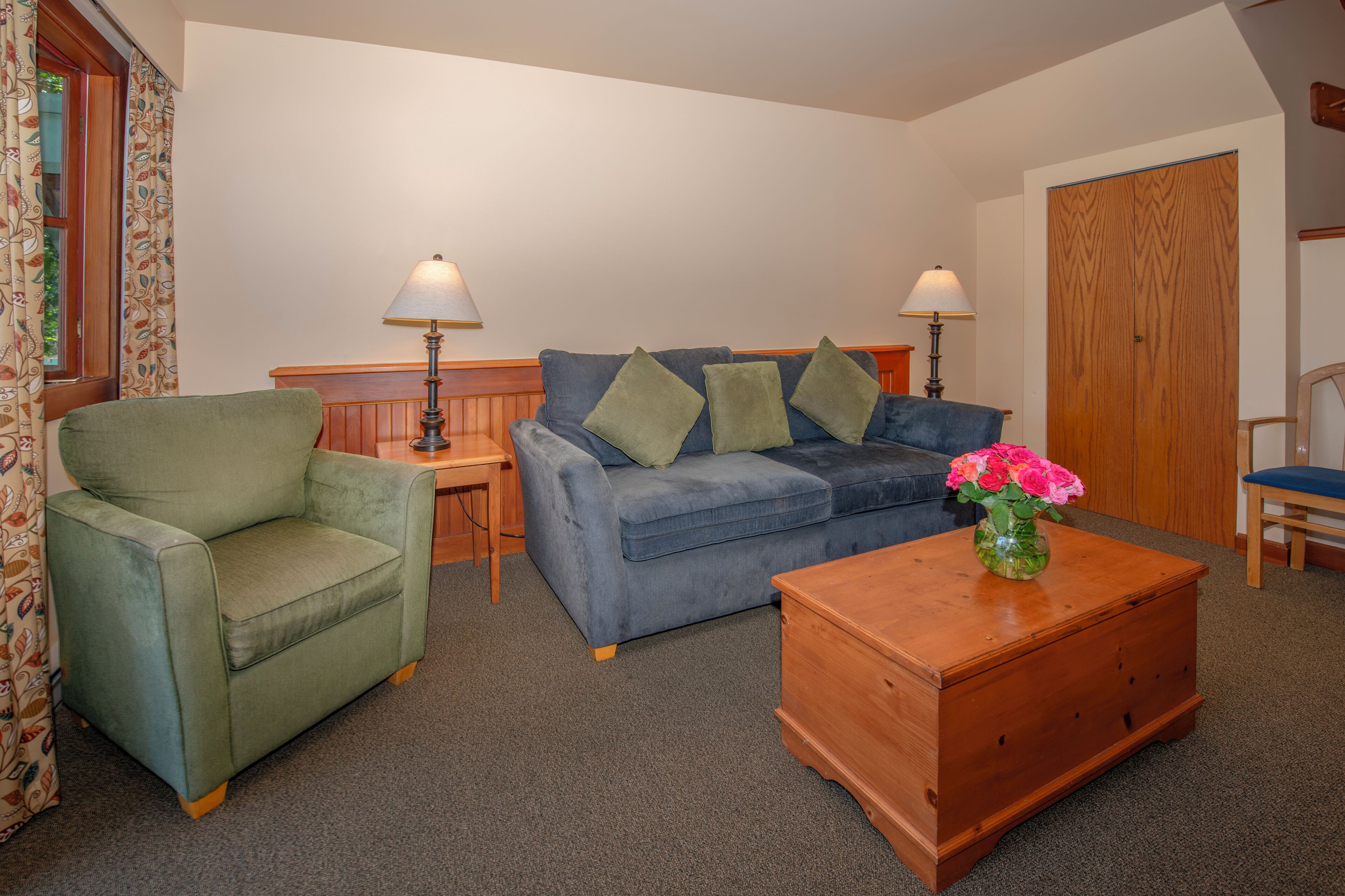 A living room with a green armchair, a blue sofa, a wooden coffee table with a vase of flowers, and two lamps on side tables.