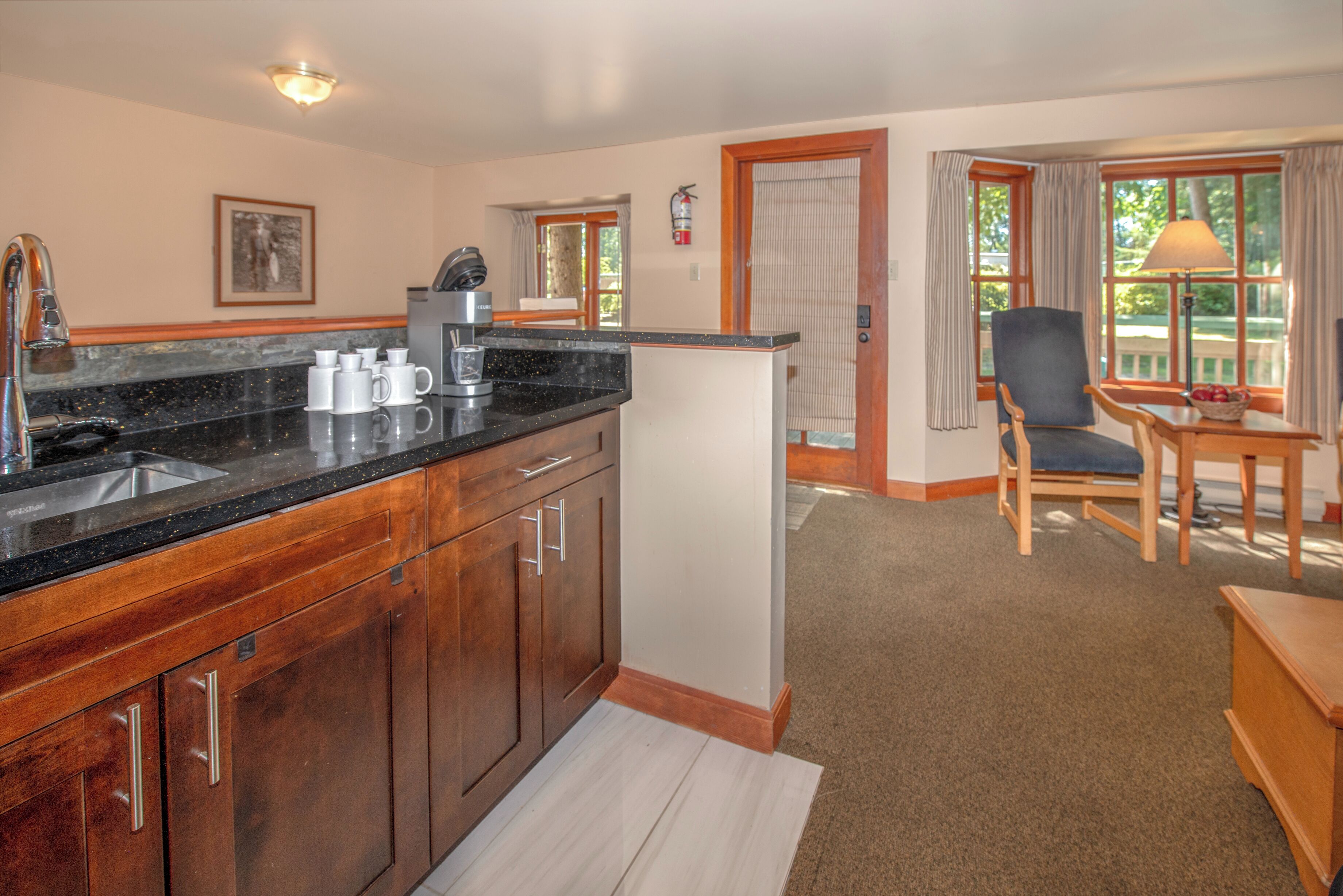 A kitchen with wooden cabinets, a black countertop, and a coffee maker. There is a chair and a table with a lamp in the living area.