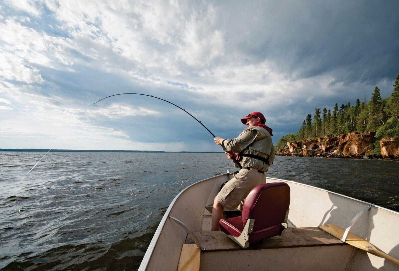 A person fishing from a small boat on a body of water with a rocky shoreline and trees in the background.