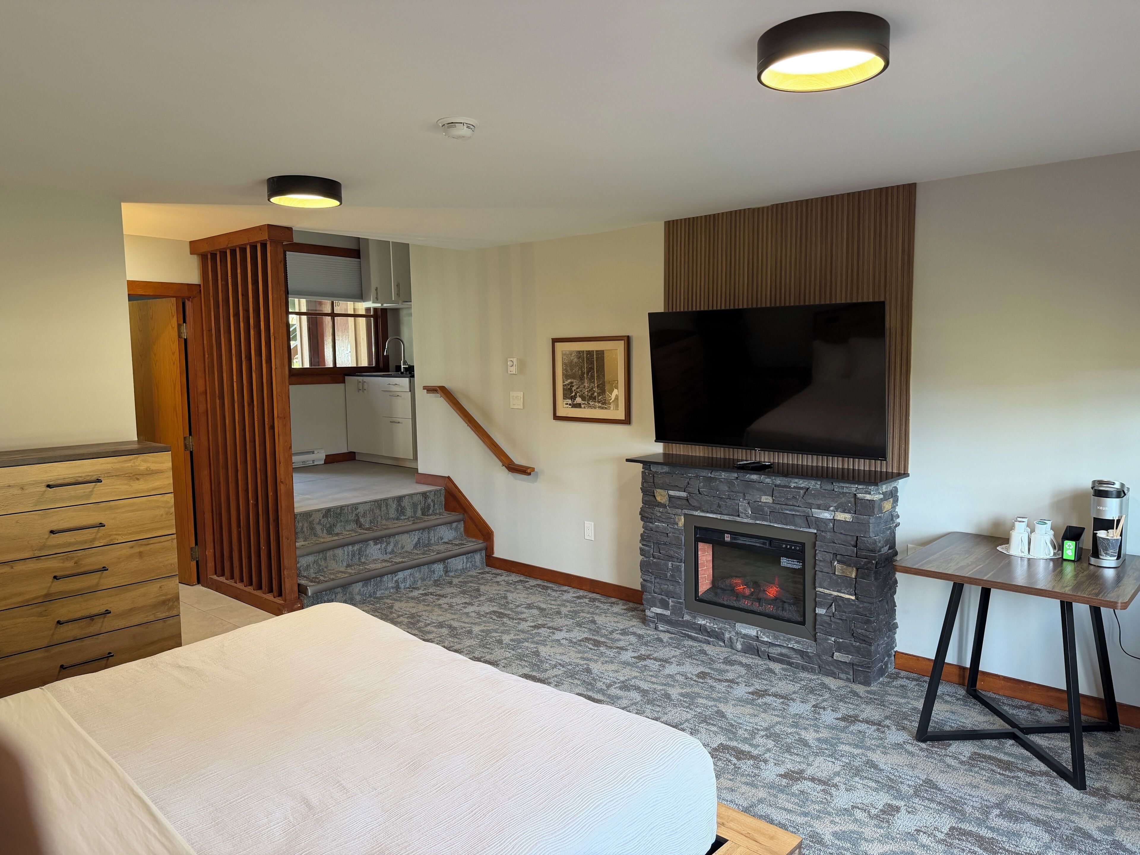 Un dormitorio con cama, televisión, chimenea, escritorio y una escalera que conduce a una cocina.