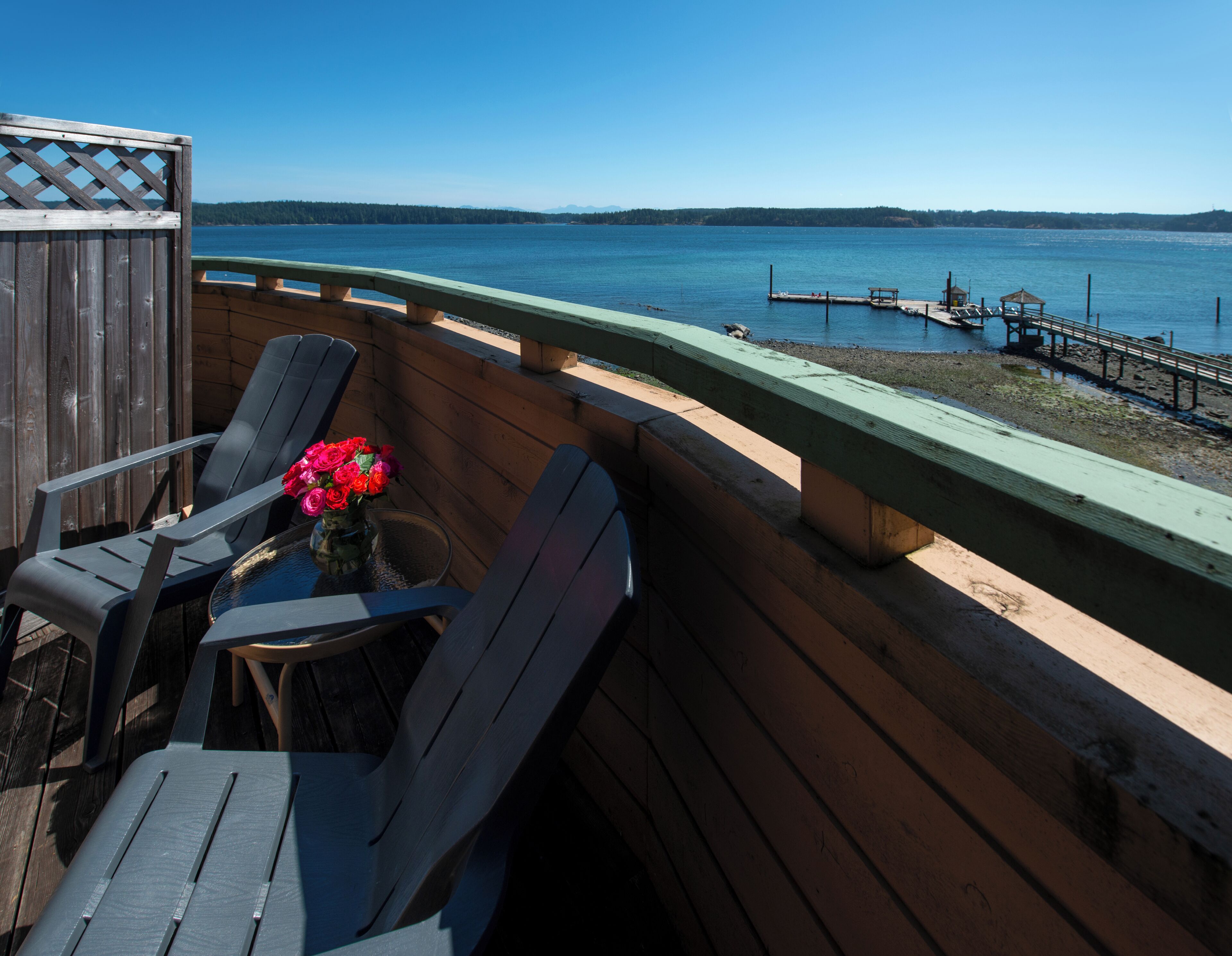 A wooden deck with a table and chairs, overlooking a calm lake with a dock and pier in the distance.