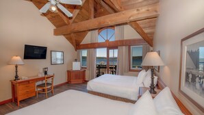 A hotel room with two beds, a desk, a chair, a TV, and a large window with a view of the sea.