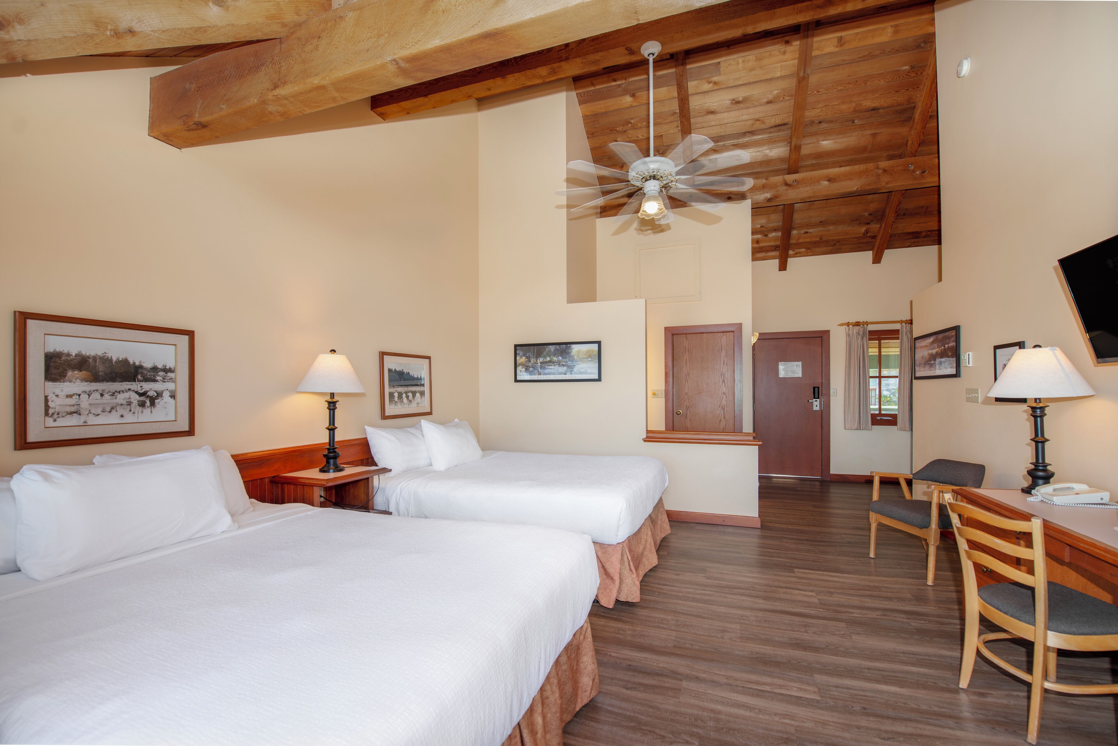 Une chambre d’hôtel avec deux lits, des poutres apparentes en bois au plafond, un ventilateur de plafond, un bureau avec une chaise et des tableaux encadrés aux murs.