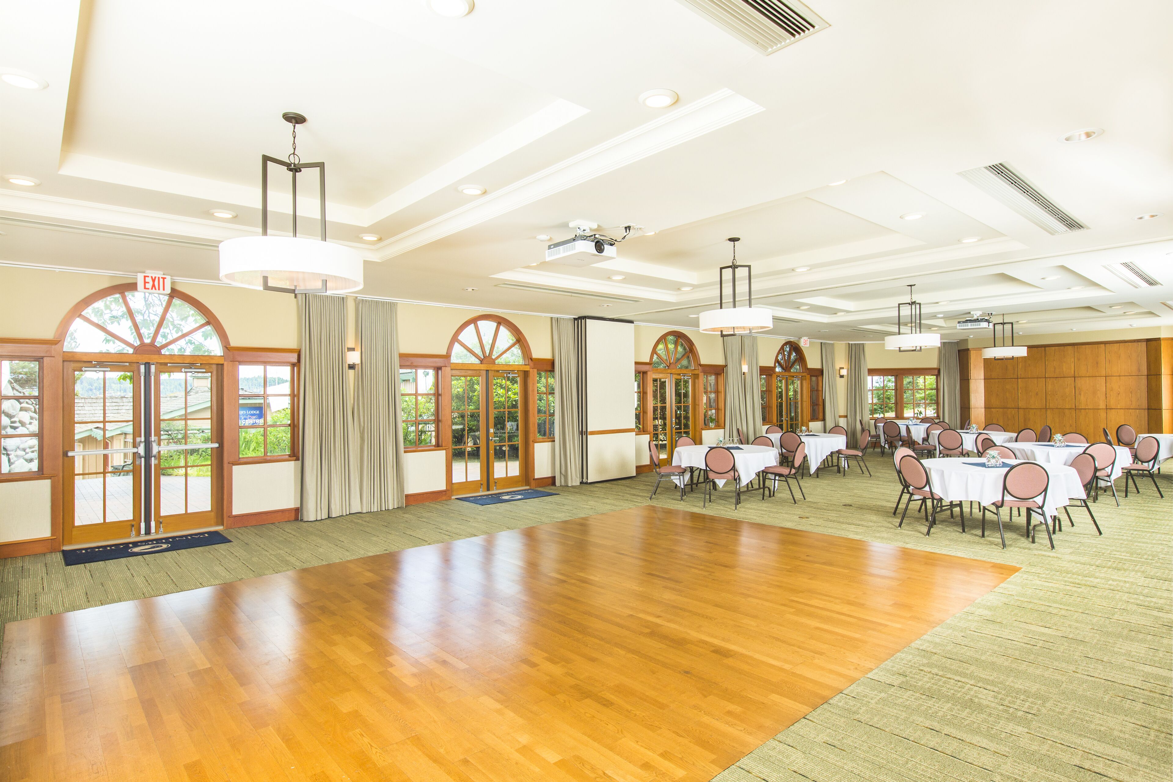 A spacious hall with wooden flooring, multiple tables with chairs, large windows, and an exit sign.