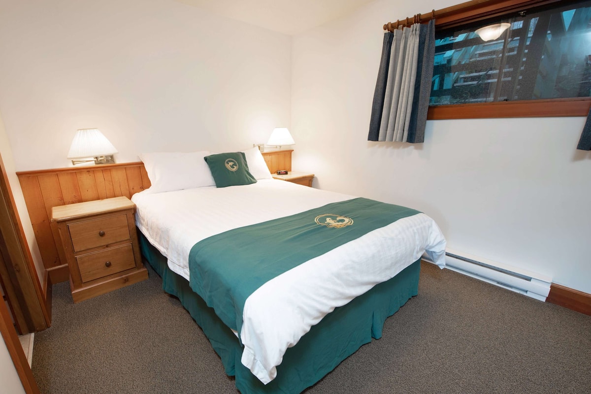 A neatly made bed with a green and white bedspread, two bedside tables with lamps, a wooden nightstand, and a window with curtains.