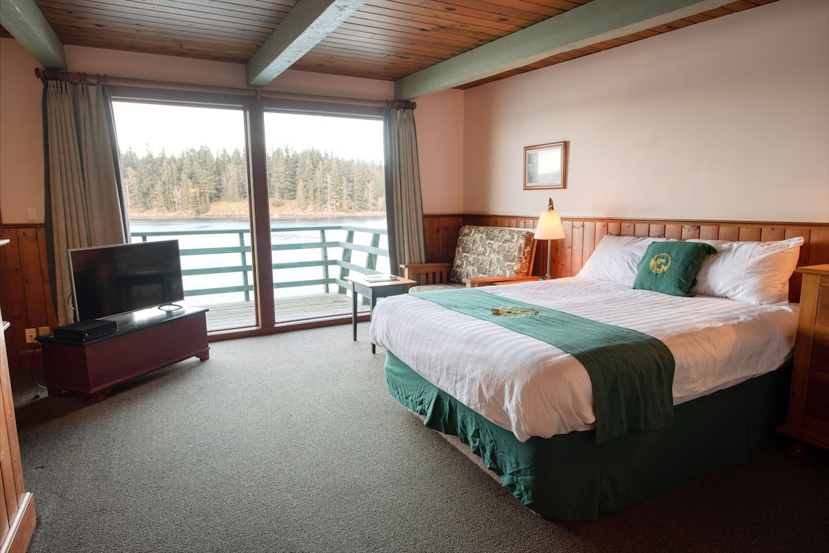 A hotel room with a bed, a television, a desk, and a view of a lake and forest.