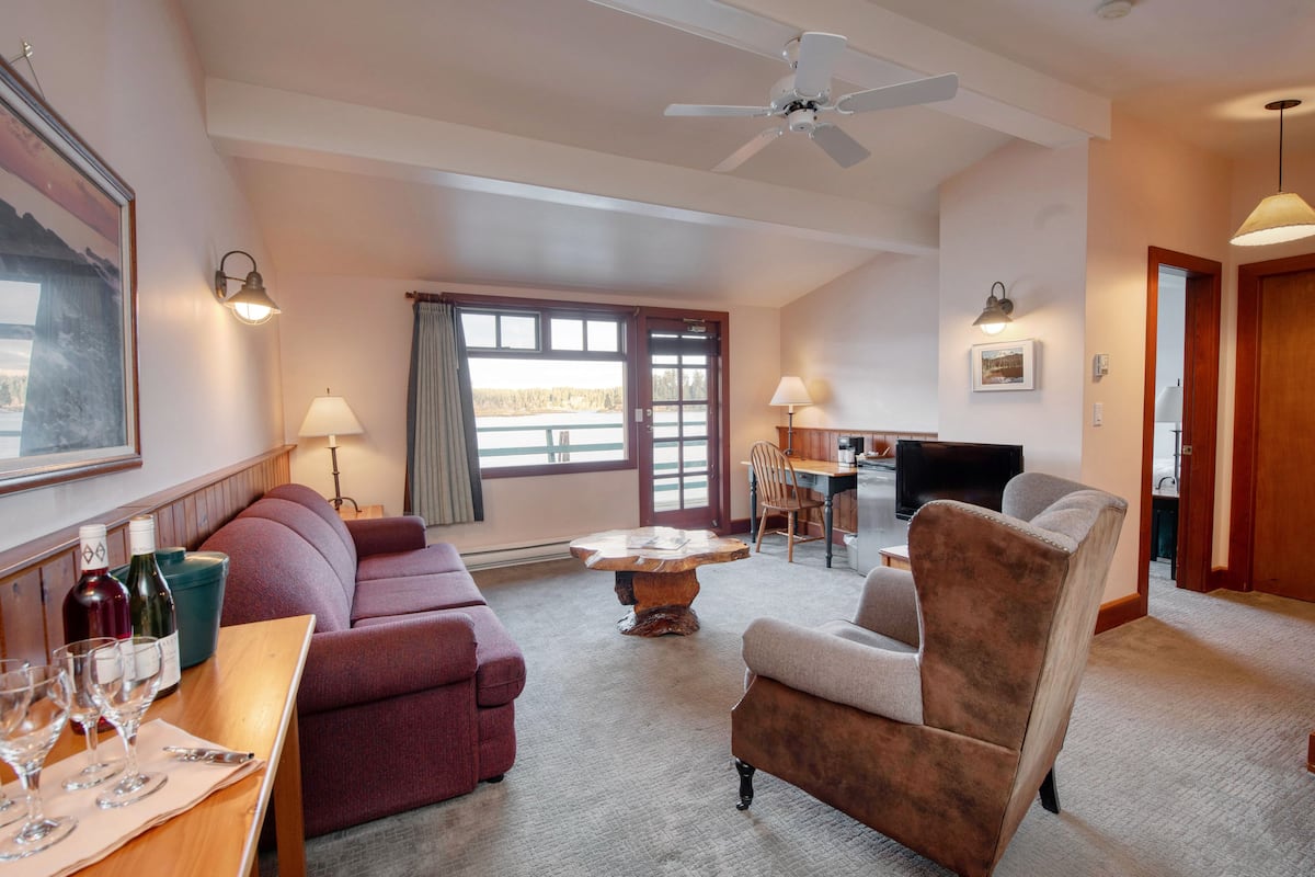 A spacious living room with a sofa, armchair, and dining table. There is a ceiling fan, a large window with curtains, and a television.