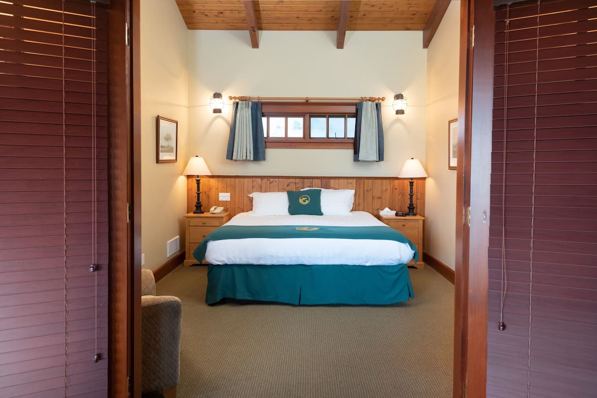 A hotel room with a wooden ceiling, a large bed with a green and white bedspread, two bedside lamps, a nightstand with a phone, and a window with curtains.