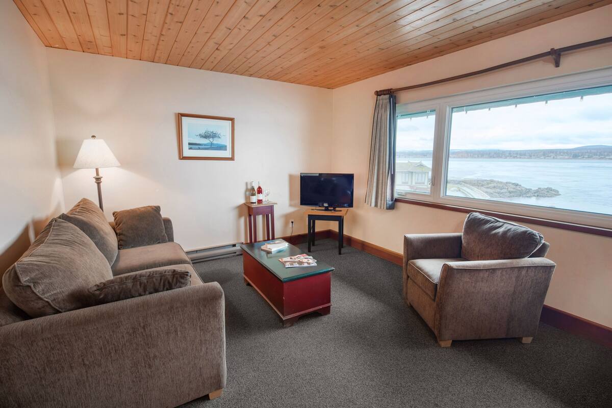 A cozy living room with a sofa, armchair, and a small coffee table. There's a TV on a stand and a window with a view of the sea.