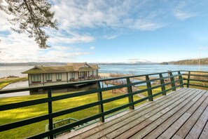 A wooden deck with a green railing overlooking a lake, a small building with a sloped roof, and distant snow-capped mountains.