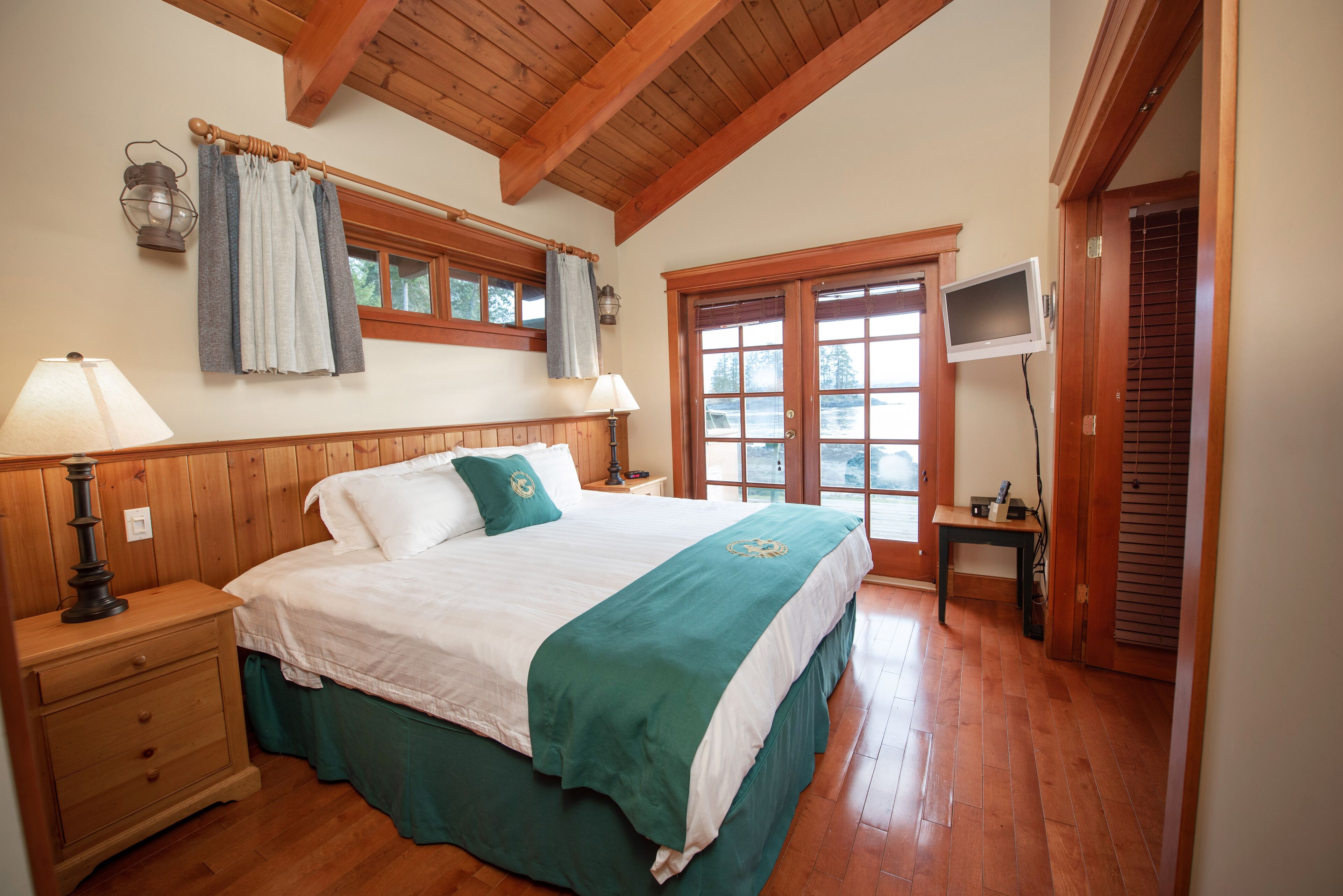 Une chambre avec un plafond en bois, un lit recouvert d’une couverture verte, deux lampes de chevet, une table de chevet, une fenêtre avec des rideaux et un téléviseur posé sur un meuble.