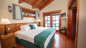 Une chambre avec un plafond en bois, un lit recouvert d’une couverture verte, deux lampes de chevet, une table de chevet, une fenêtre avec des rideaux et un téléviseur posé sur un meuble.
