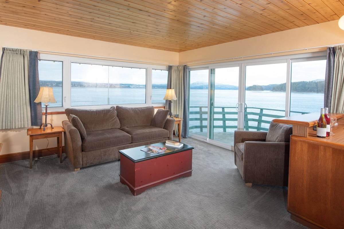 A cozy living room with a sofa, armchair, and coffee table, large windows with a view of a body of water, and a wooden ceiling.