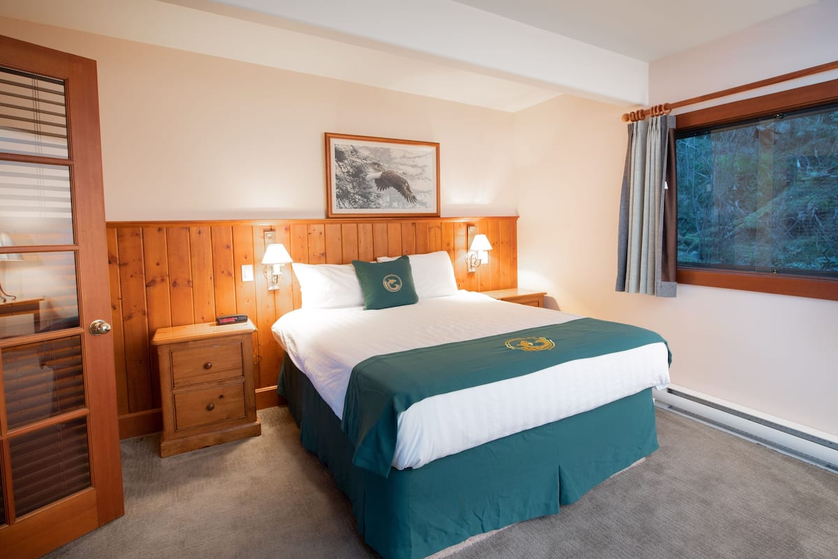 A hotel room with a wooden headboard, a large bed with a teal and white bedspread, two bedside lamps, a nightstand, and a window with curtains.