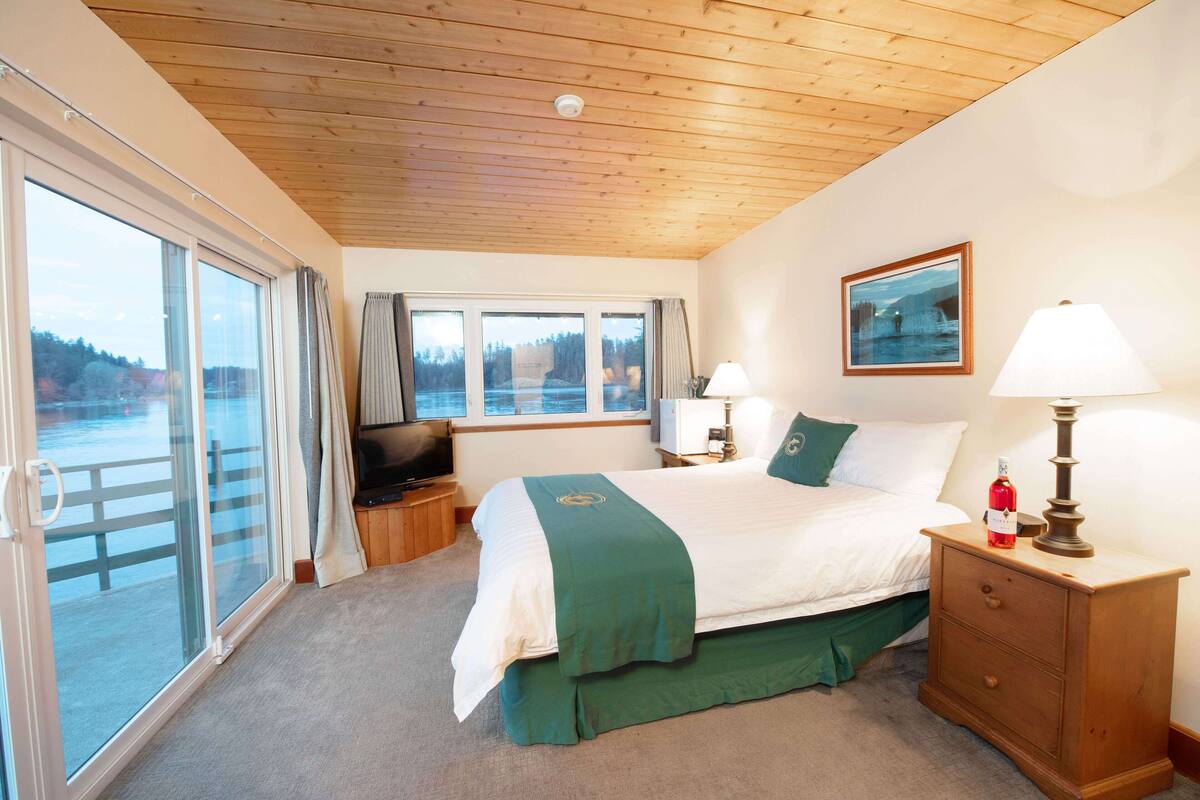 A hotel room with a large bed, a wooden ceiling, a window with a view of the outdoors, a painting on the wall, and a nightstand with a lamp.