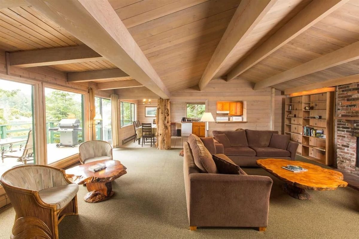 A spacious living area with a wooden ceiling, large windows, a fireplace, and a cozy seating arrangement.