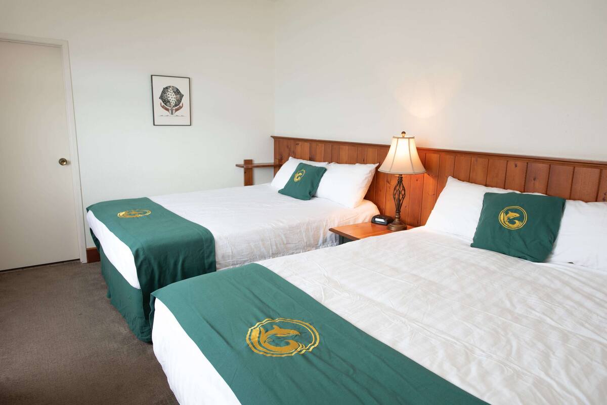 A hotel room with two beds, each with a green bedspread featuring a logo, white linens, and a wooden headboard.
