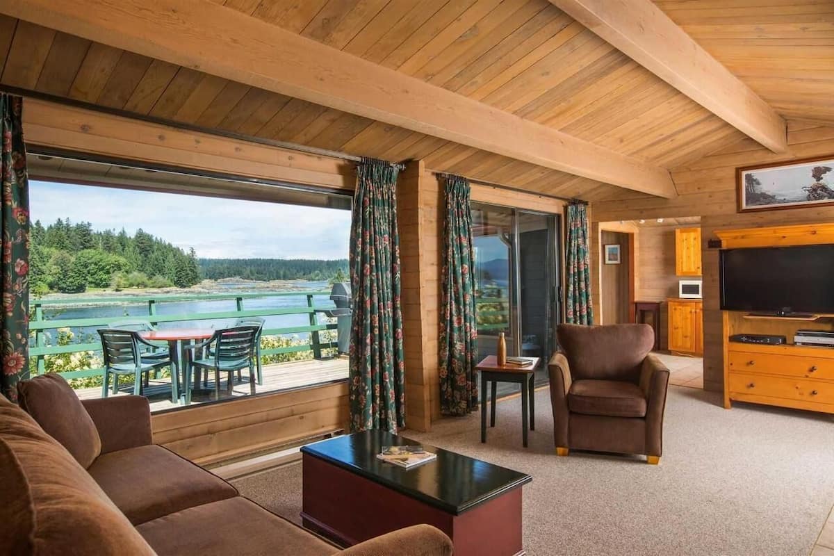 A cozy living room with a large window overlooking a scenic lake, wooden furniture, and a flat-screen TV.