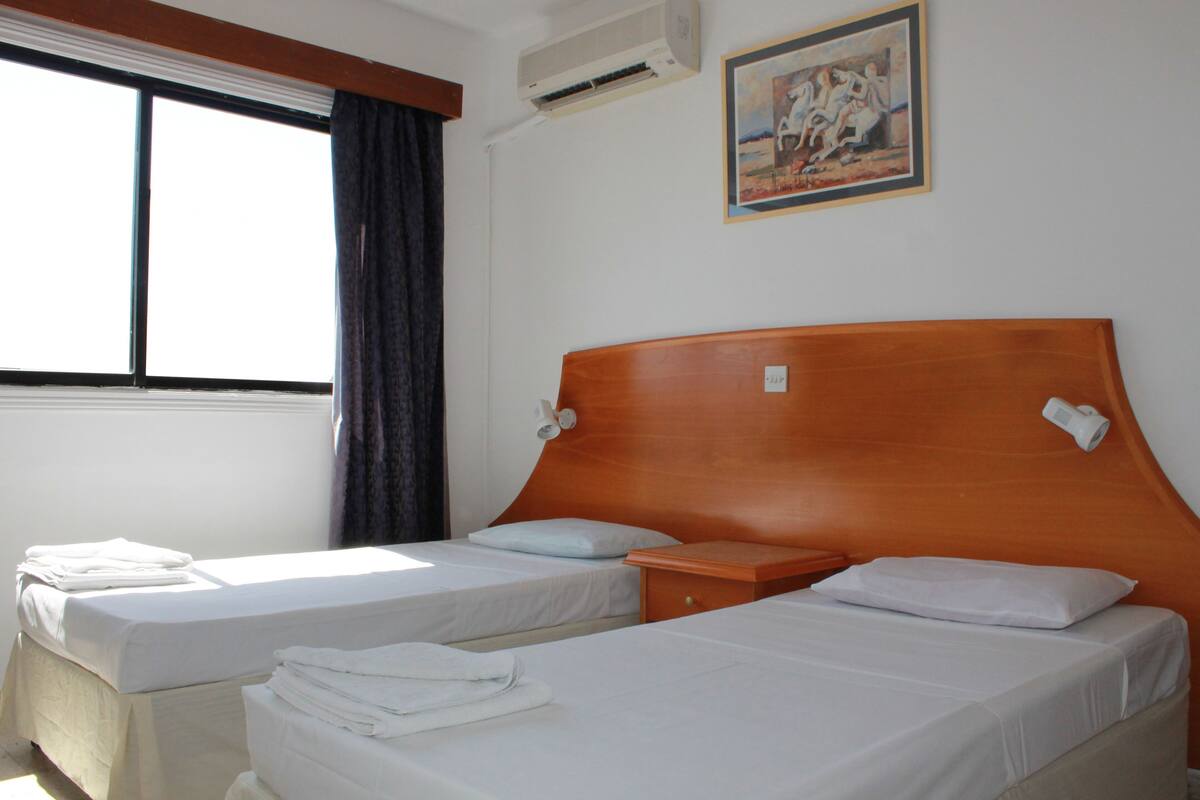 A hotel room with two single beds, a wooden headboard, a small bedside table, and a painting on the wall.