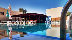 Une piscine avec des chaises longues et des parasols, un espace de détente couvert et un ciel d’un bleu limpide.
