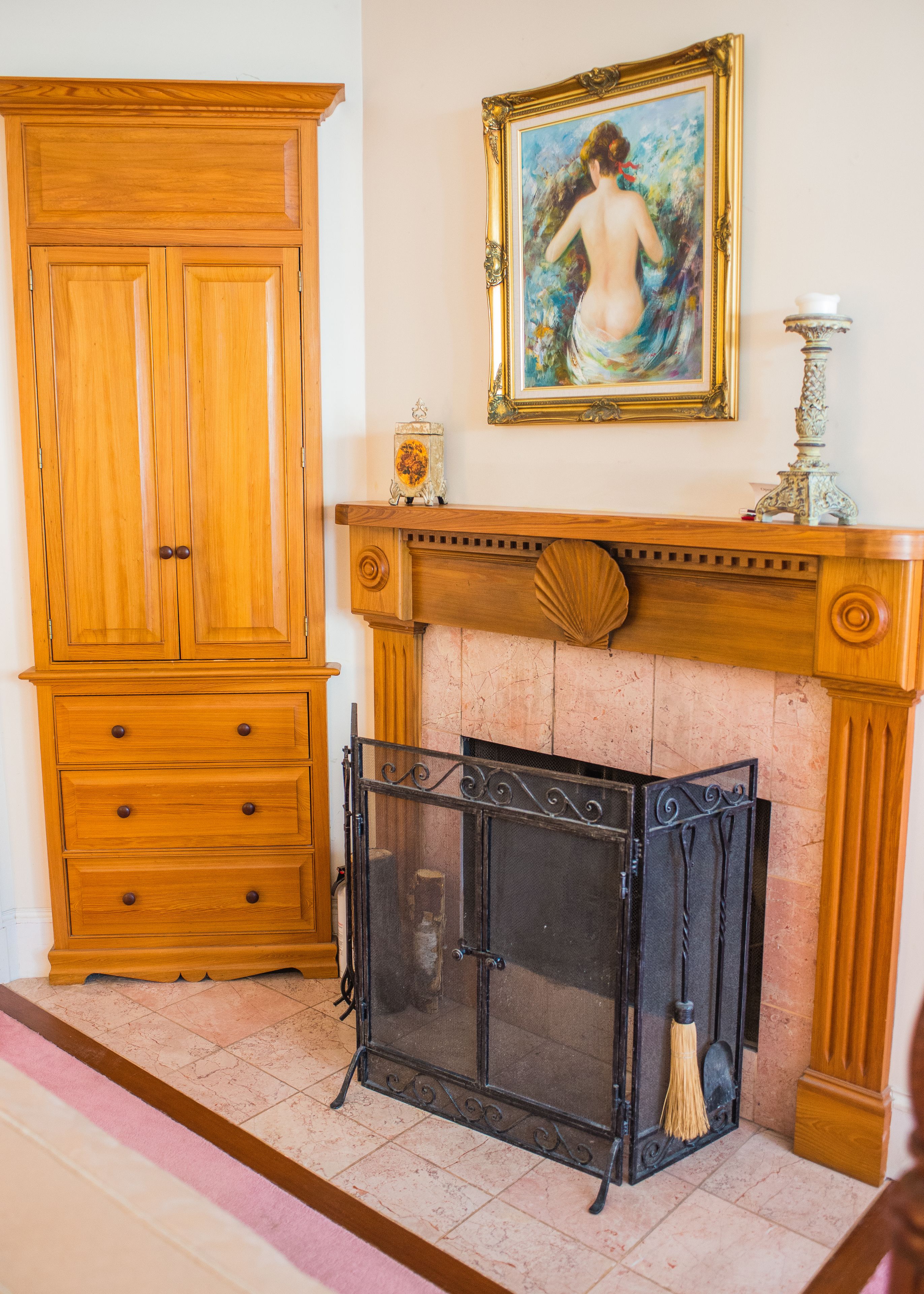 A room with a wooden wardrobe, a fireplace with a screen, and a framed painting on the wall.