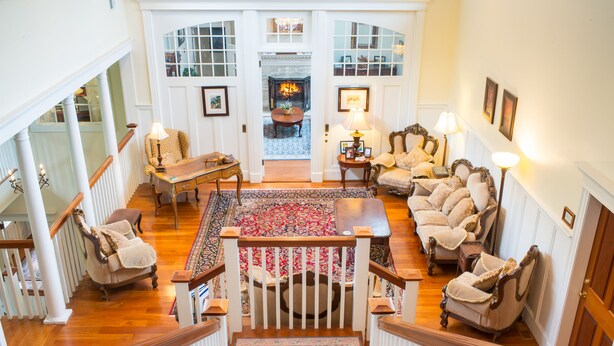 A spacious living room with a high ceiling, a grand staircase, and a fireplace. - Chiltern Inn (Bar Harbor)