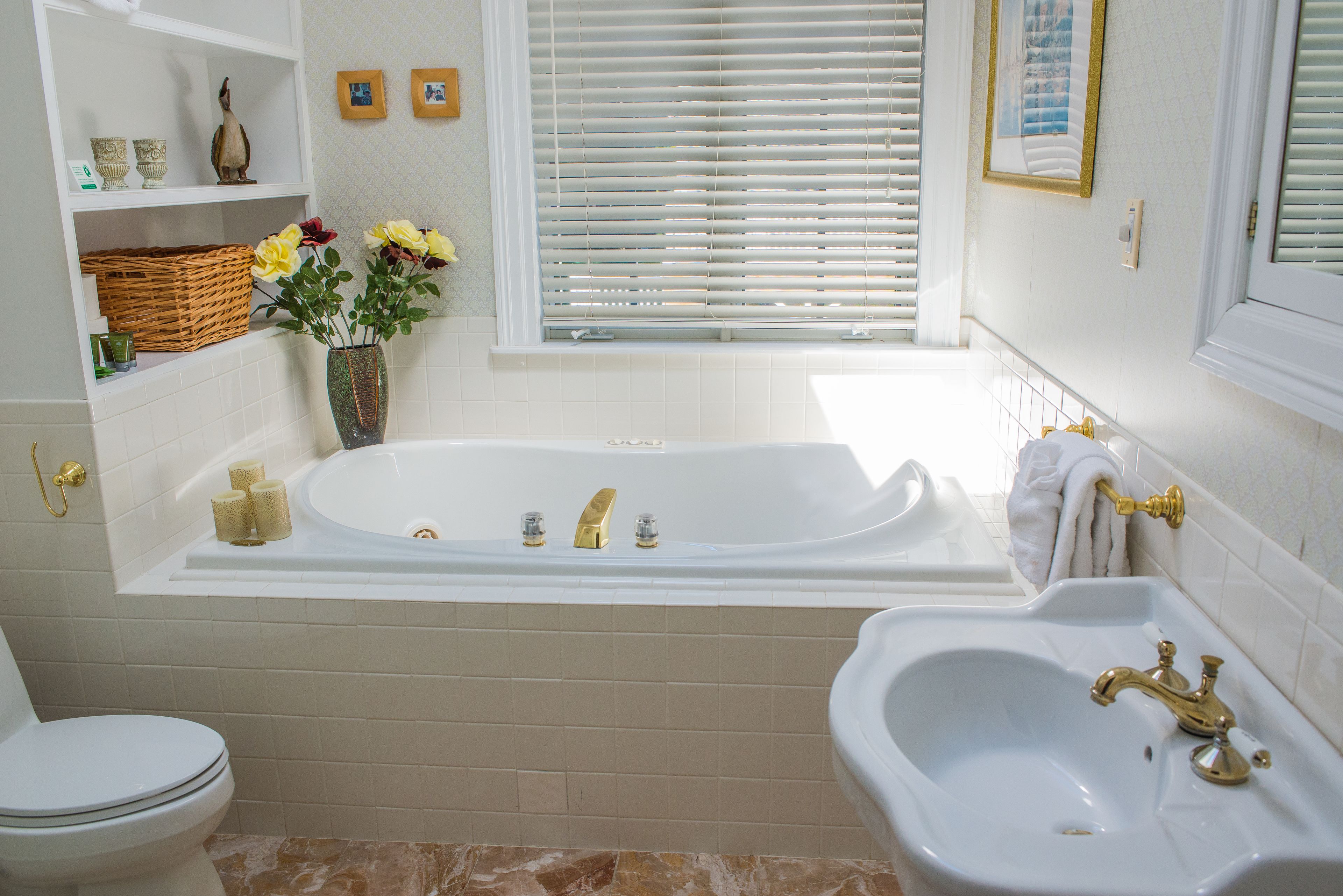 Een badkamer met een bad, een wastafel en een toilet. Er staan planken met decoratieve voorwerpen en een raam met jaloezieën.