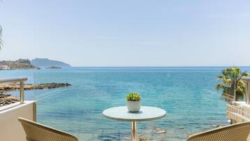 A balcony with a round table and two chairs, overlooking a clear blue sea and a rocky shoreline.