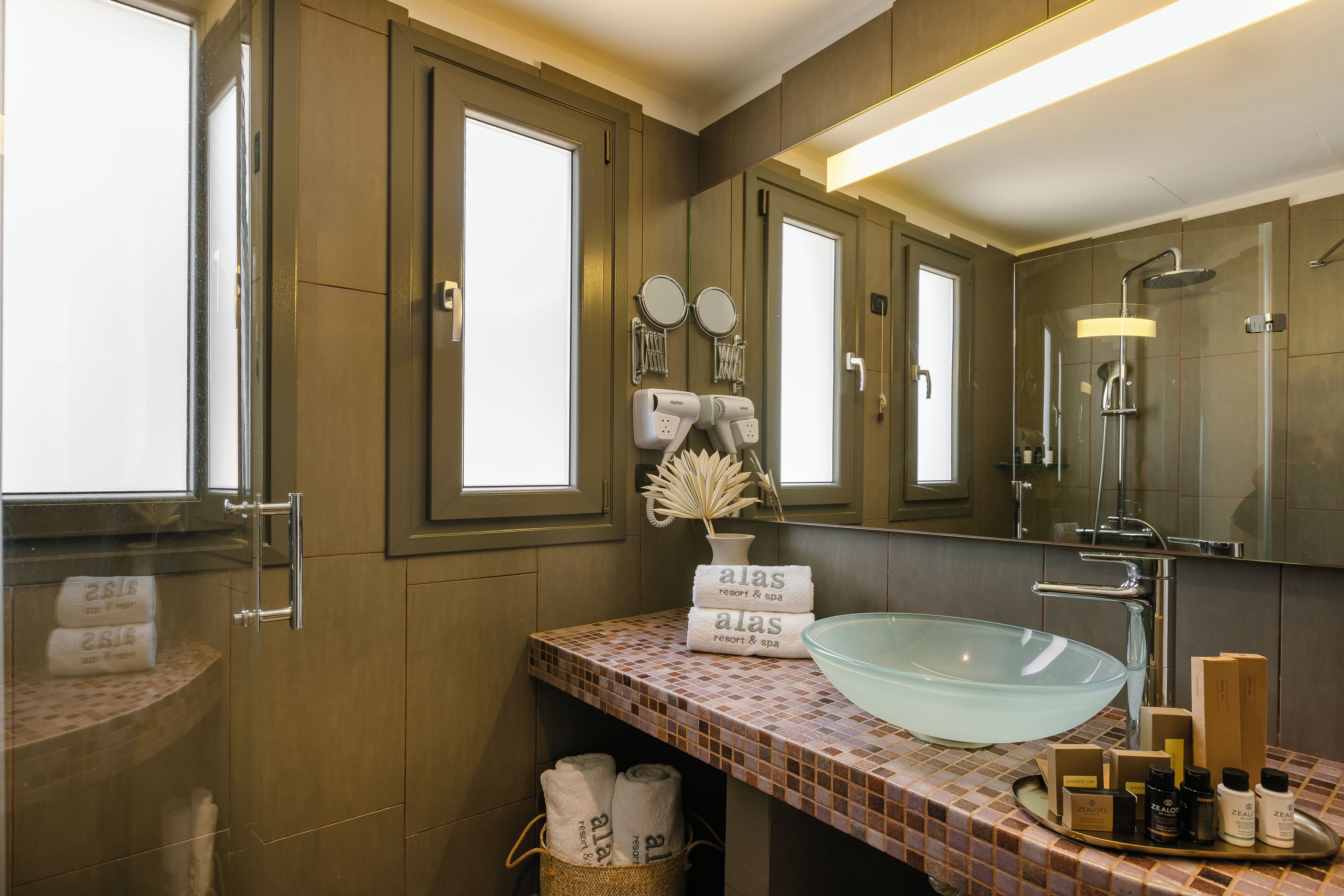 A bathroom with a large glass bowl sink, a mirror, and a shower. There are towels and toiletries on the counter.