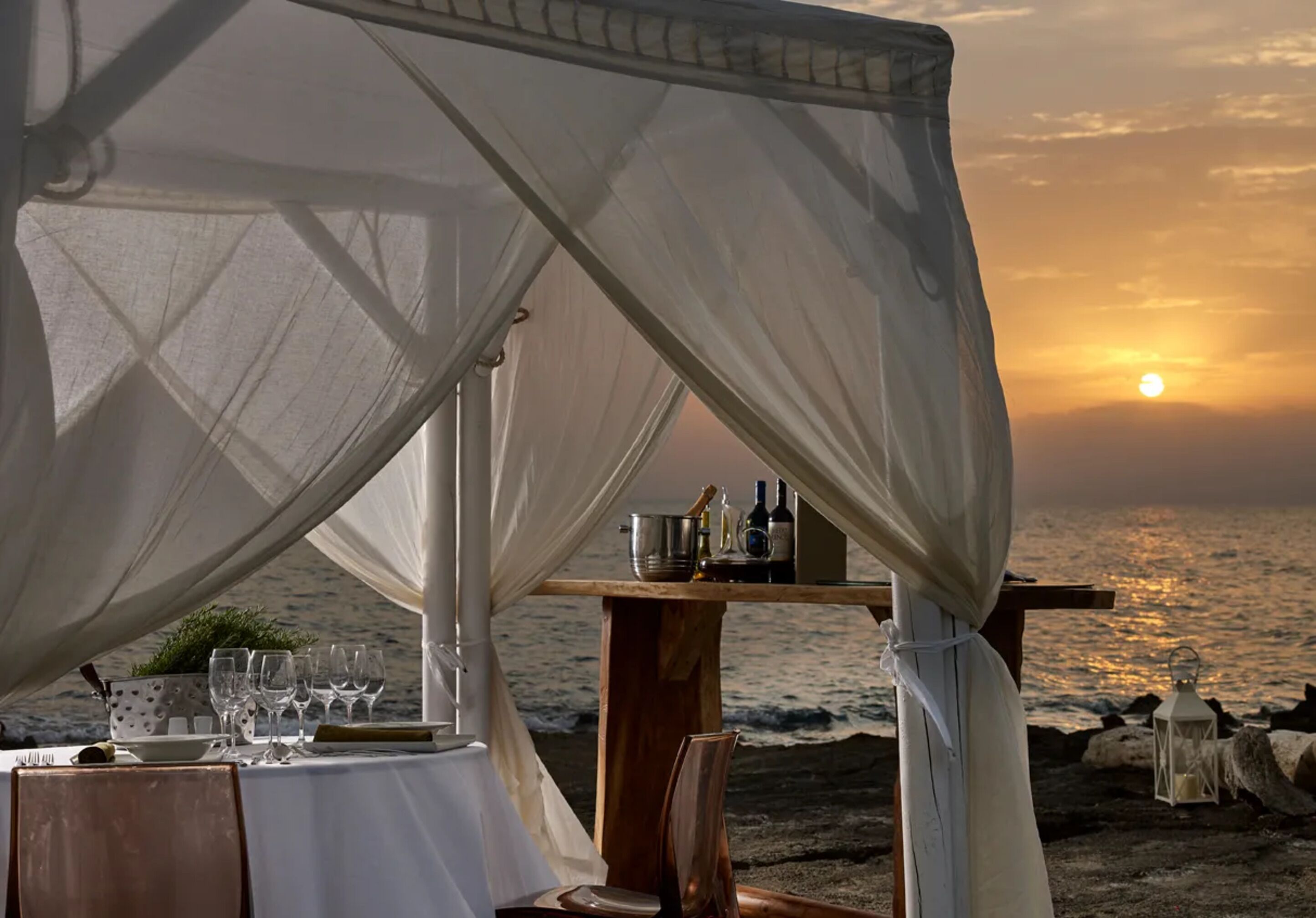 Un montaje para cenar frente al mar, con una mesa dispuesta para dos personas, con vista al océano al atardecer.