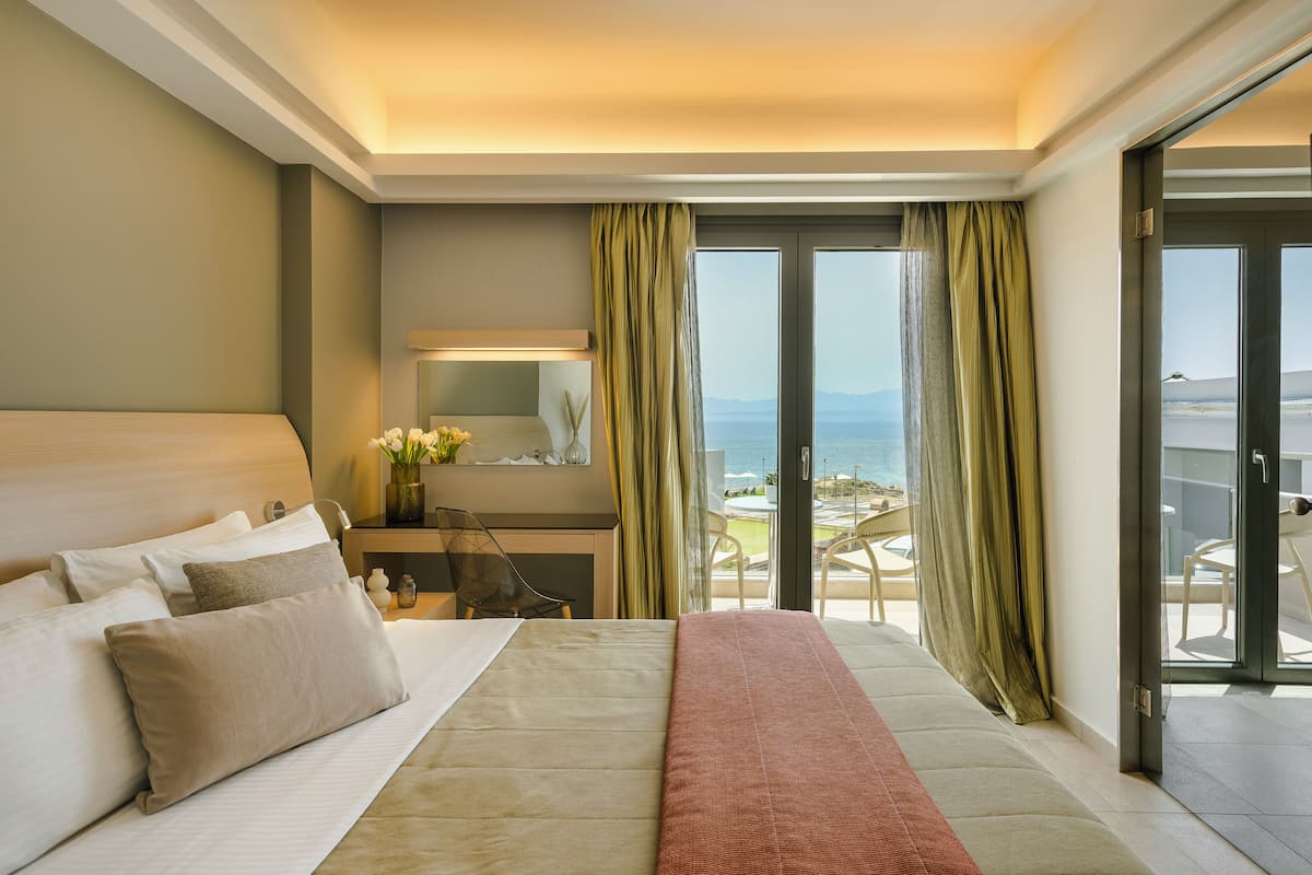 A hotel room with a large bed, a desk, a chair, and a view of the beach through the sliding glass door.