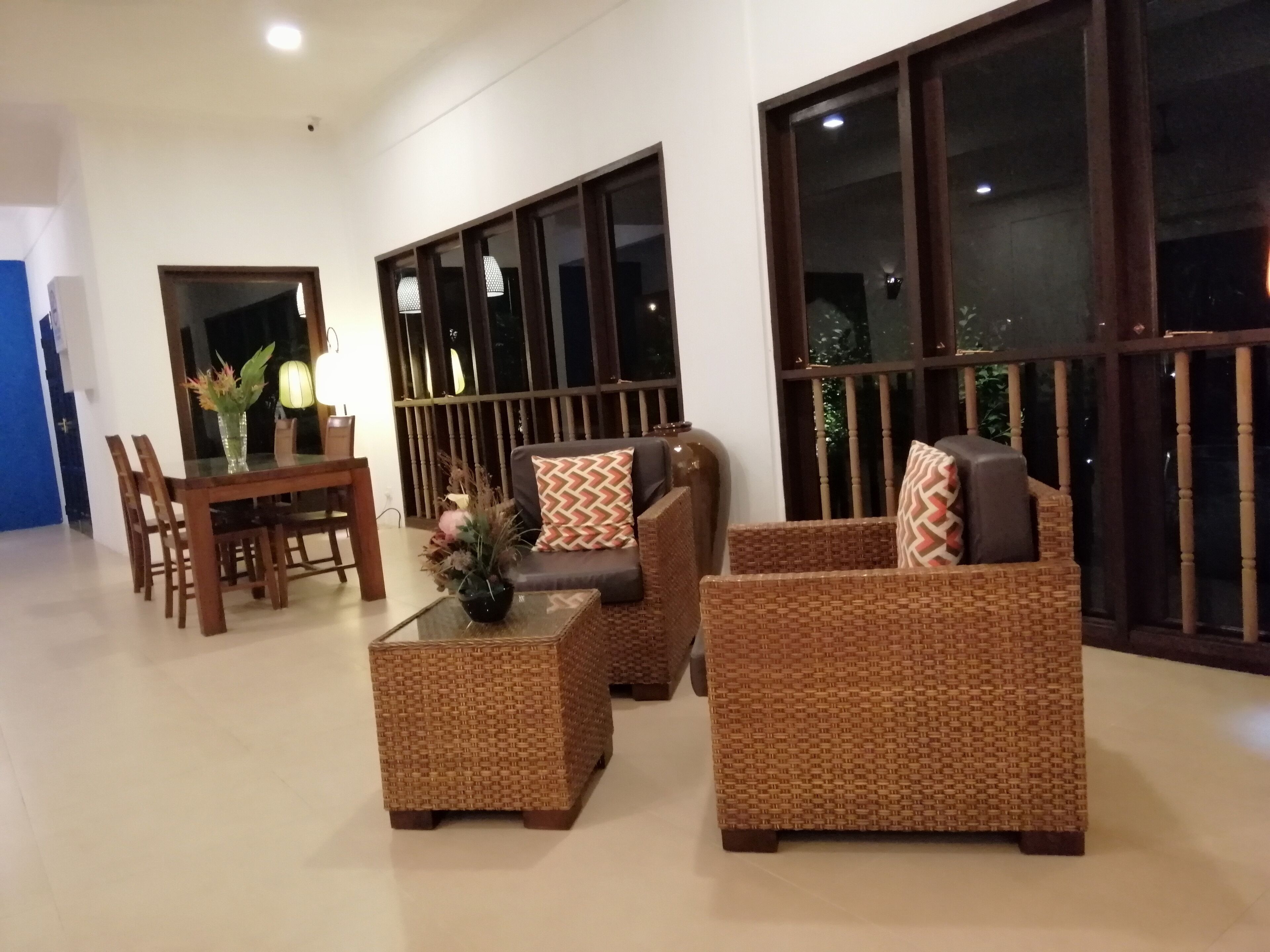 A modern living room with a dining area, featuring wicker furniture, a glass coffee table, and large windows with dark frames.