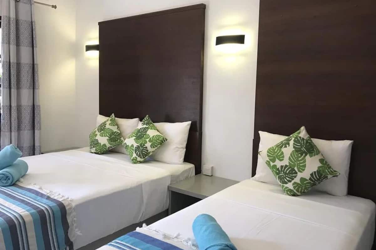 A hotel room with two single beds, each with a dark headboard and a white bedspread. There are two green leaf-patterned pillows on each bed. The room has a curtain on the left side and a wall-mounted light fixture above the beds.