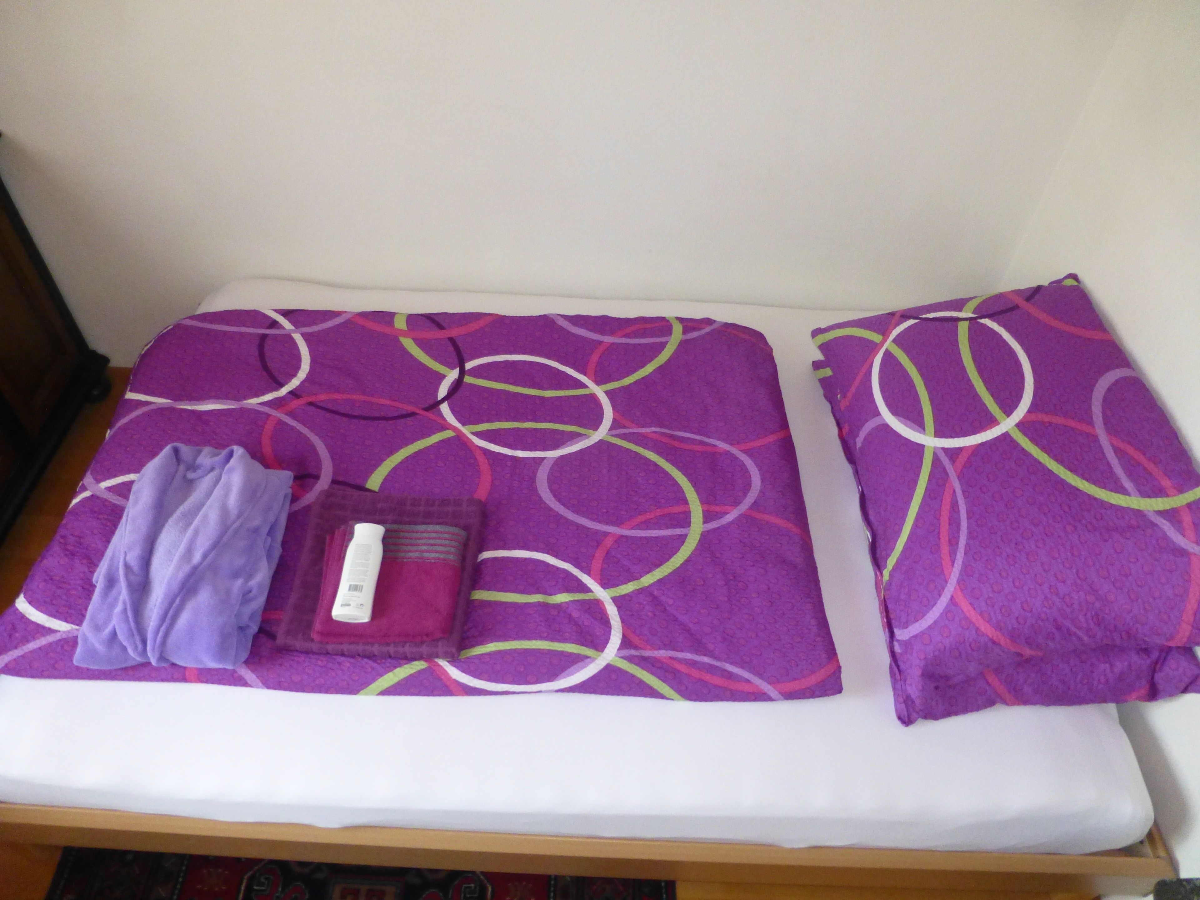 A small bed with a purple and white patterned bedspread, a folded purple towel, and a white pillow.