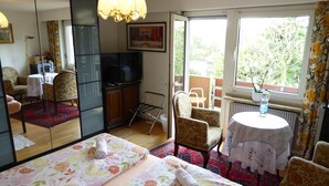 A hotel room with a bed, a TV, a small table, a chair, and a balcony with a view of trees. - B&B Pappelweg - 2 (Muttenz)