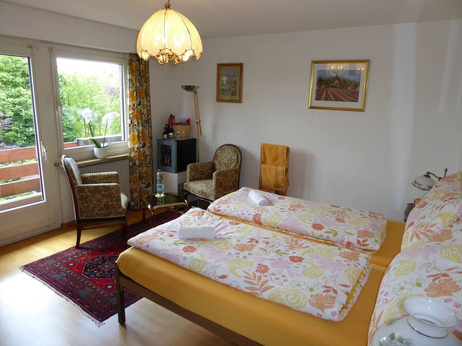 A bedroom with a bed, two chairs, a table, a radiator, a window with curtains, and a hanging light fixture.