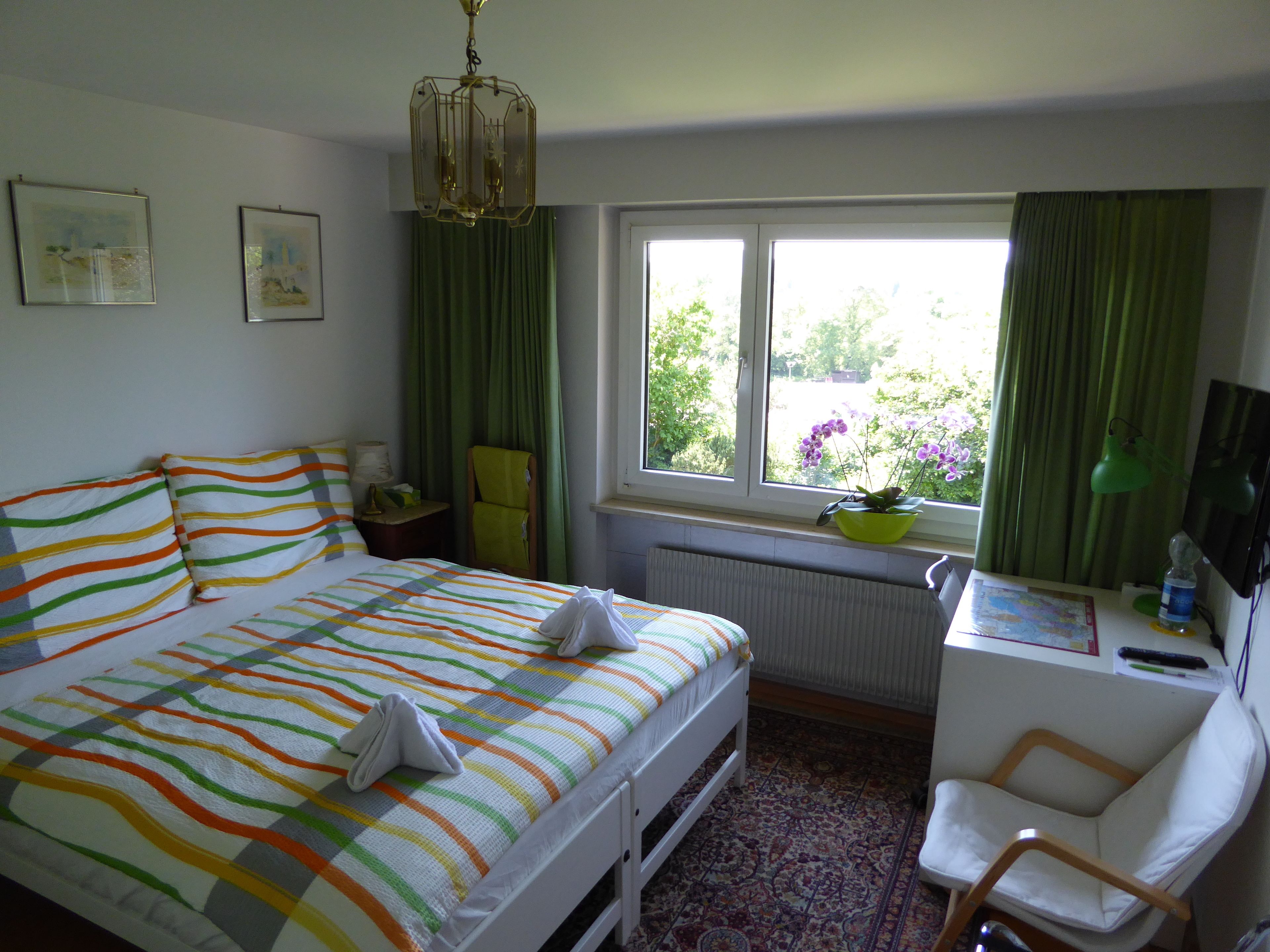 A bedroom with a bed, a chair, a desk, a window with curtains, and a hanging light fixture.