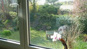 A view through a window of a garden with a bench, a small table, and a flowering plant. - B&B Pappelweg - 2 (Muttenz)
