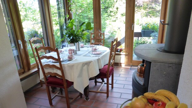 A dining area with a round table, chairs, a bowl of fruit, and a view of greenery through large windows. - B&B Pappelweg - 2 (Muttenz)