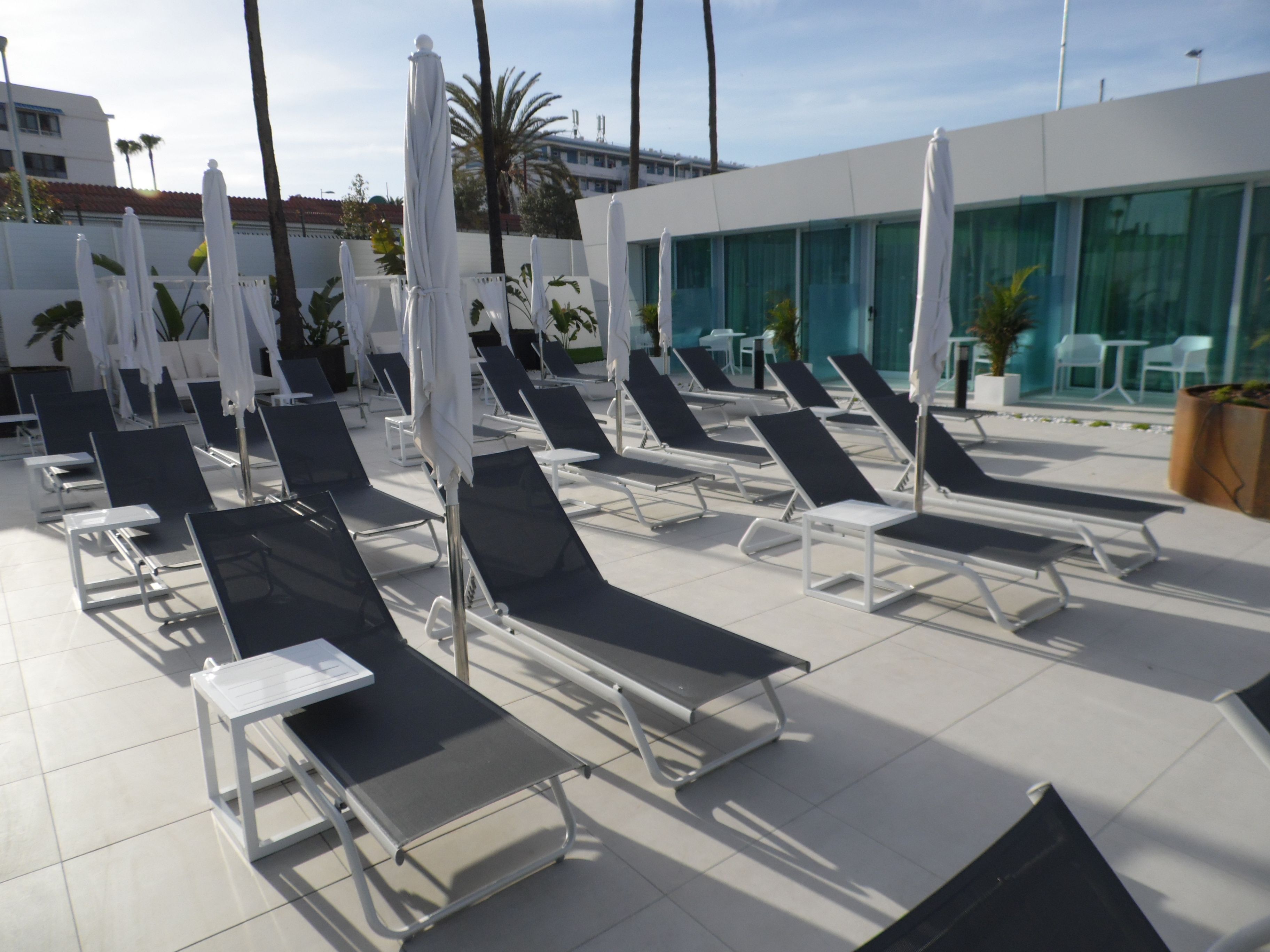 Área de piscina com espreguiçadeiras, guarda-sóis e um edifício moderno ao fundo.