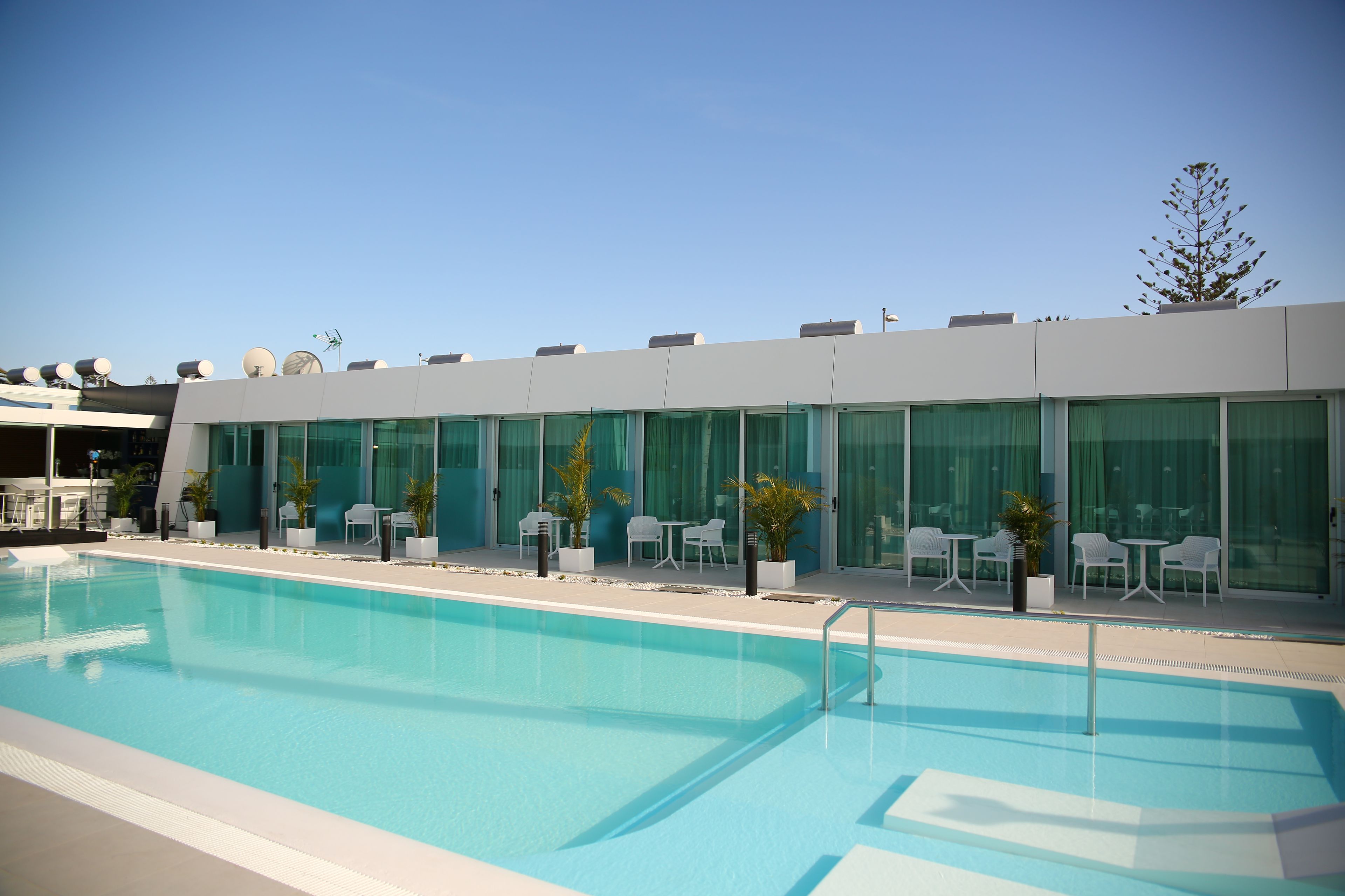 A modern hotel with a swimming pool, outdoor seating, and a clear sky.