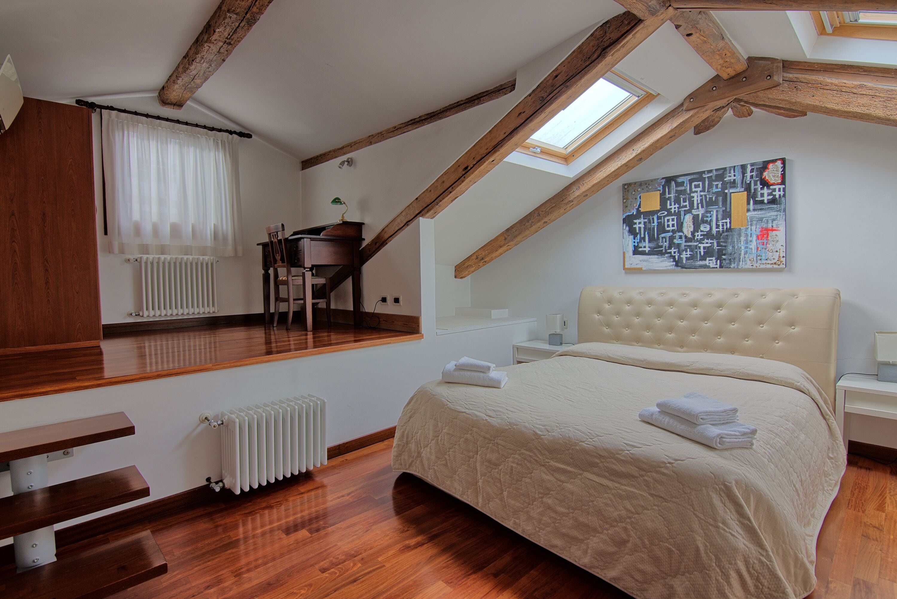A bedroom with a sloped ceiling, wooden beams, a large bed, a radiator, a window with curtains, a bedside table, and a wall-mounted artwork.