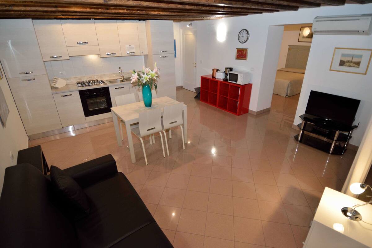 A modern living room with a black sofa, a dining area with a table and chairs, a kitchen with white cabinets, and a red entertainment unit.