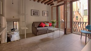 Ein Wohnzimmer mit Sofa, Couchtisch und Sessel. Ein Balkon mit Geländer bietet Blick auf umliegende Gebäude.