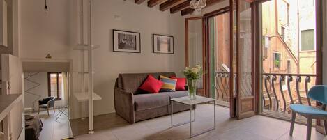 Un soggiorno con un divano, un tavolino e una poltrona. C'è un balcone con ringhiera e vista sugli edifici.