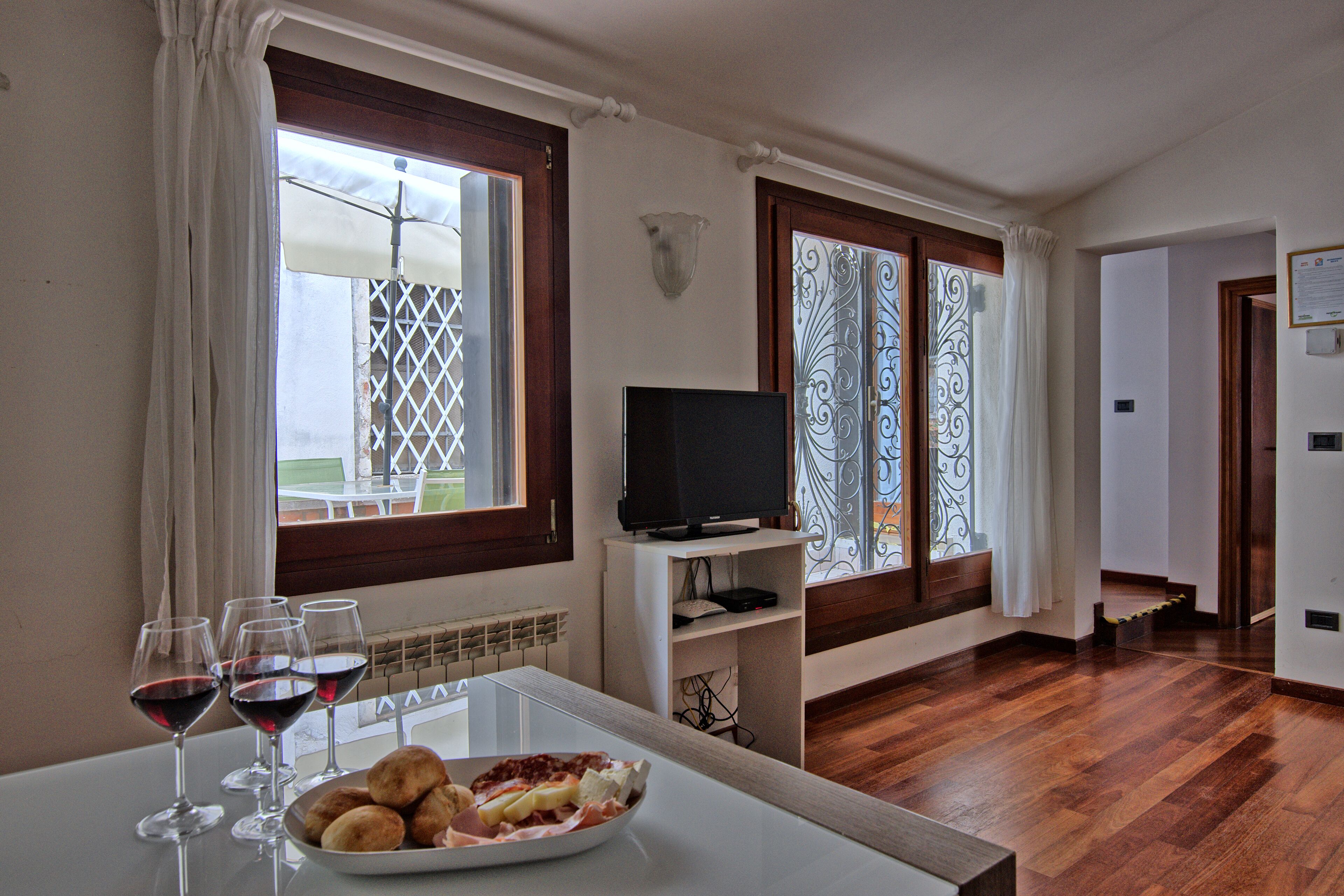 A room with a dining table set with wine glasses and a plate of food, a TV on a stand, and large windows with decorative iron curtains.